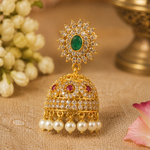An ornate gold-toned jhumka earring with a green oval stone at the top, surrounded by clear stones, a dome-shaped lower section with red and clear stones, and five white pearl drops, displayed on a beige fabric surface with blurred flowers and a brass vessel in the background.