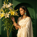 A woman in a cream-colored saree stands beside a large bouquet of white and yellow lilies, wearing ornate silver-toned earrings with turquoise accents, set against a dark green arched wall.
