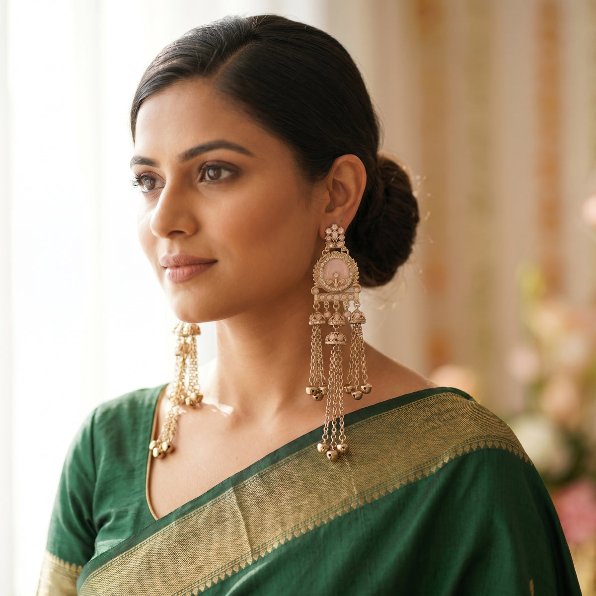 A woman wearing ornate gold-toned traditional Indian earrings with circular motifs and dangling chains, paired with a green saree, shown in a softly lit indoor setting.