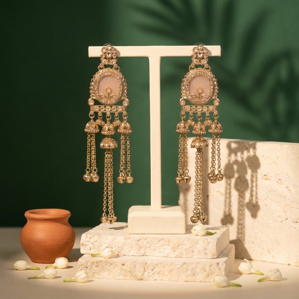 A pair of ornate gold-toned dangle earrings with oval pale pink stone centers and dangling chain bells, displayed on a white T-stand against a dark green backdrop with decorative props.