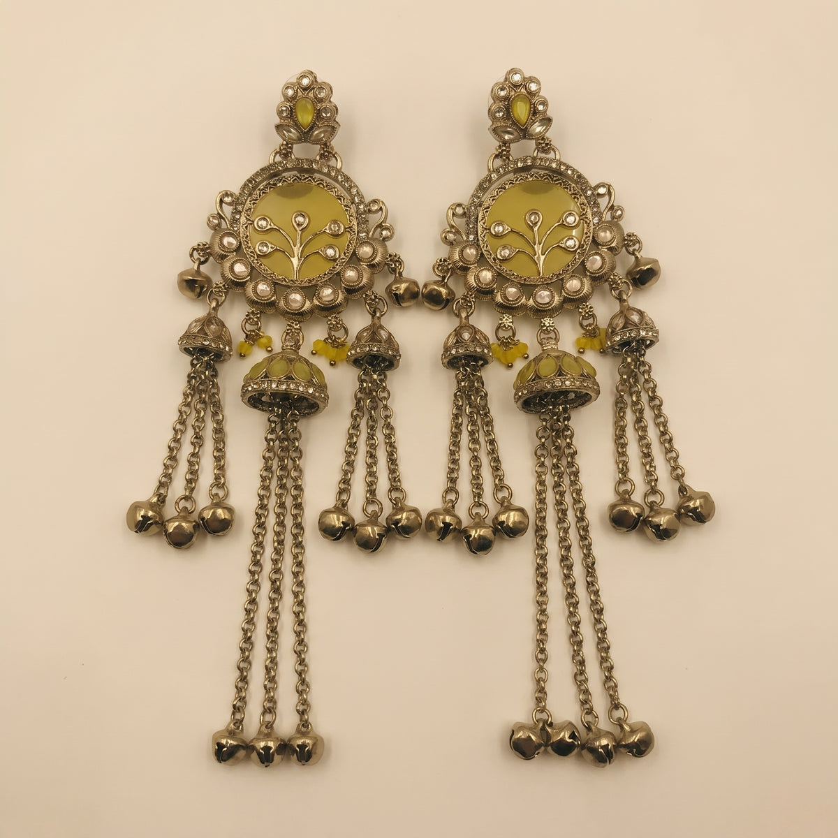 A pair of ornate silver-toned Indian chandelier earrings with yellow-green enamel inlays, floral motifs, and dangling bell charms, displayed on a plain beige background.