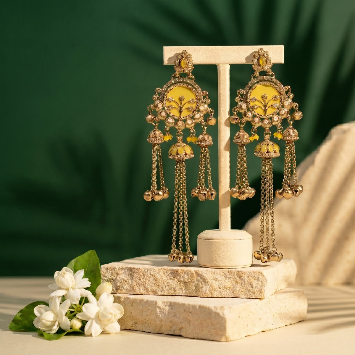 A pair of ornate gold-toned dangle earrings with yellow enamel panels and dangling chain bells, displayed on a cream jewelry stand. The stand is placed on stacked stone blocks with white flowers at the side, against a dark green leafy backdrop.