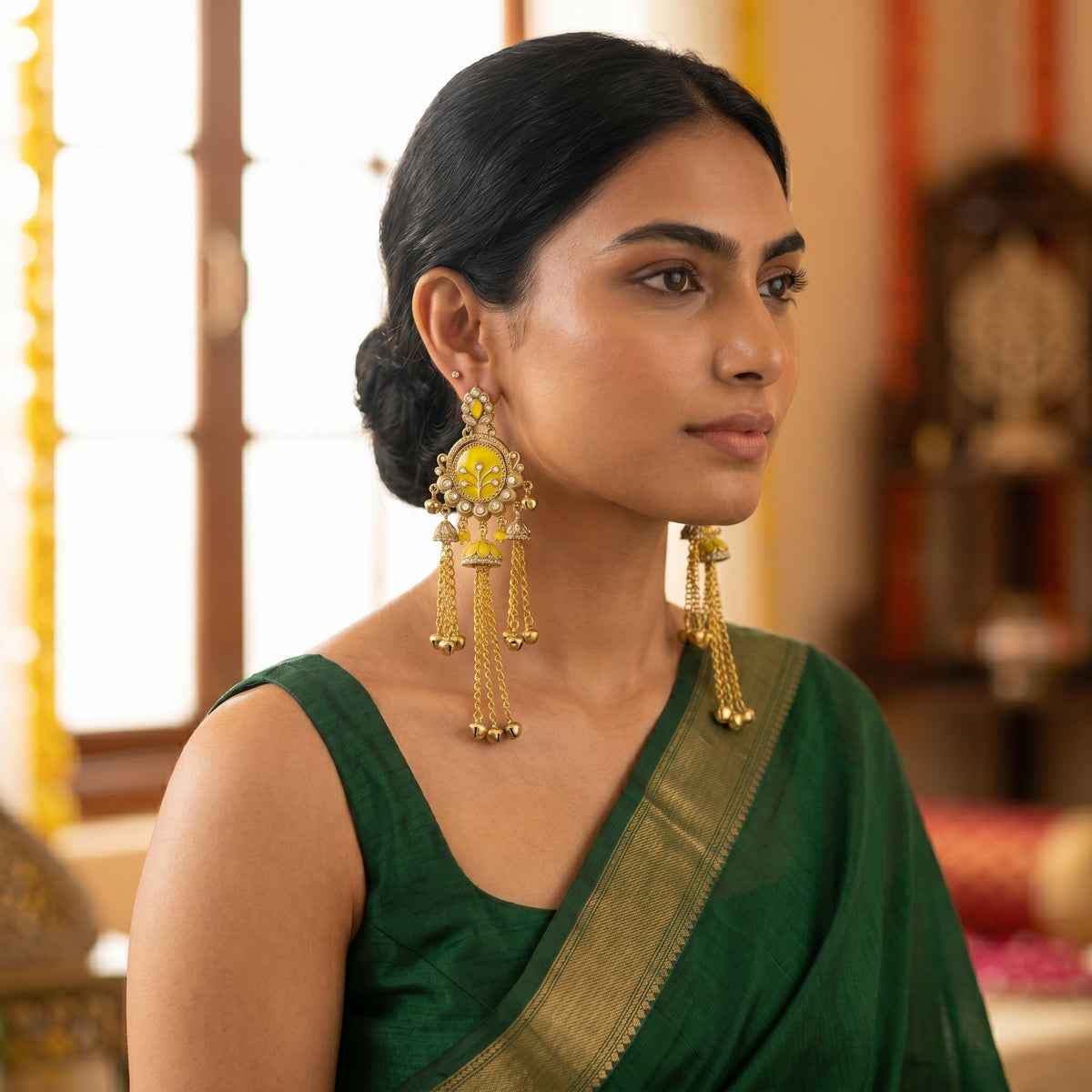 A woman wearing a dark green saree and large gold-toned traditional earrings with yellow stone accents, photographed indoors with a softly blurred warm-toned background.