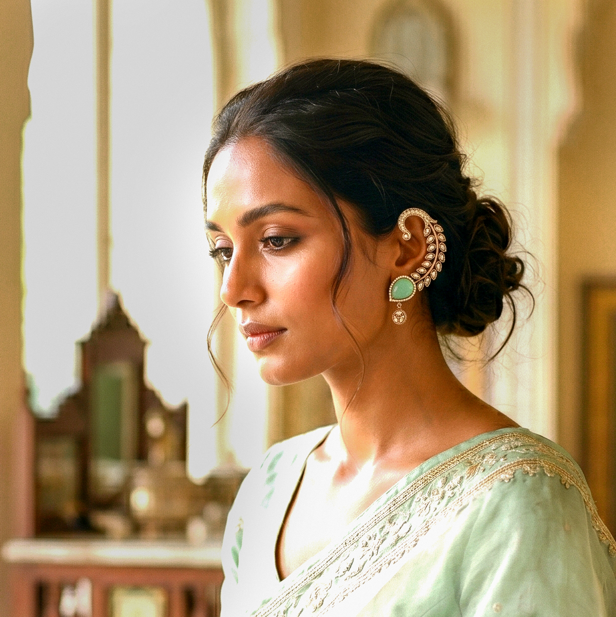 A woman in profile wearing a mint green saree and a large gold-toned traditional Indian earring with a green stone, set against a softly blurred warm interior background.