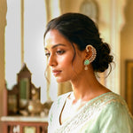 A woman in profile wearing a mint green embroidered saree and a large gold-toned traditional Indian earring with a green stone, set against a softly blurred indoor background.