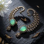 A pair of ornate gold-toned crescent-shaped earrings with large green teardrop gemstones and rows of clear stones, arranged on a dark textured surface with dried leaves and a white flower.