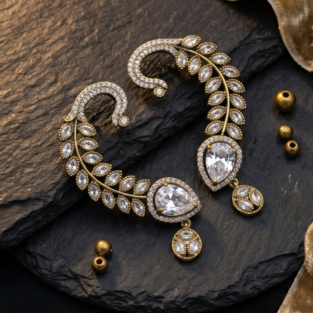Gold-toned statement earrings with clear crystal-like stones, arranged on a dark textured stone surface with small gold beads nearby.