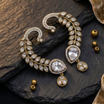 Gold-toned statement earrings with clear crystal-like stones, arranged on a dark textured stone surface with small gold beads nearby.