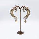 A pair of ornate gold-toned drop earrings with clear rhinestone detailing and teardrop-shaped clear stones, displayed on a decorative bronze stand against a white background.