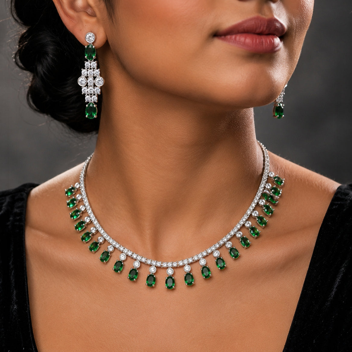 A close-up of a model wearing a silver-toned necklace and matching drop earrings set with emerald-cut green stones and clear stones, shown against a dark gray background.