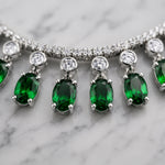 A close-up of a silver-toned tennis necklace with oval-cut emerald green gemstones and round clear stones, displayed on a white marble surface.