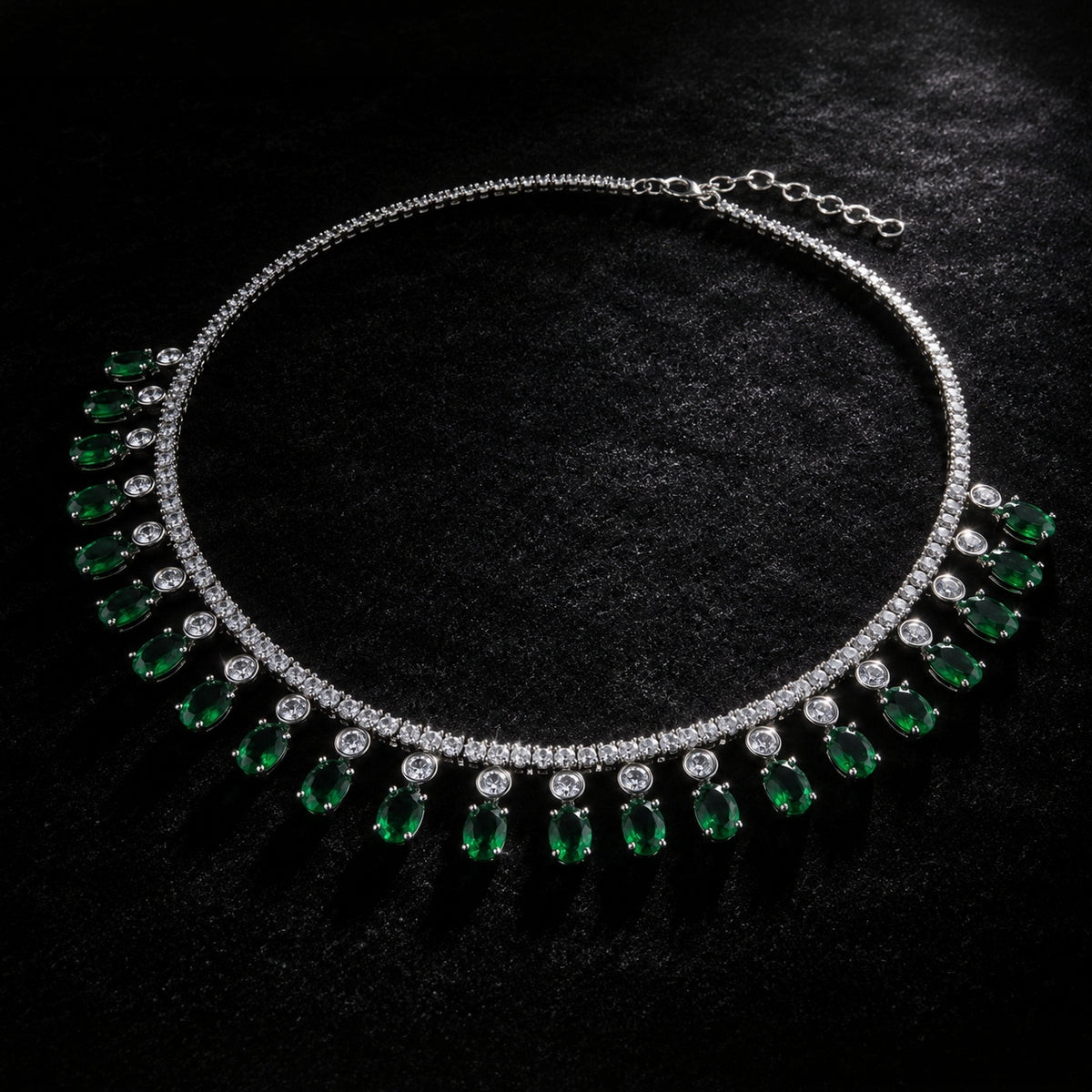 A silver-tone collar necklace featuring a row of round clear stones and a series of dangling green teardrop-shaped gemstones, displayed on a textured black background.
