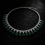 A silver-tone collar necklace featuring a row of round clear stones and a series of dangling green teardrop-shaped gemstones, displayed on a textured black background.
