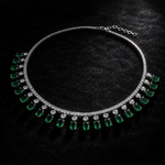 A silver-tone necklace featuring a row of green teardrop-shaped emerald-cut gemstones and clear round-cut stones, displayed on a textured black background.