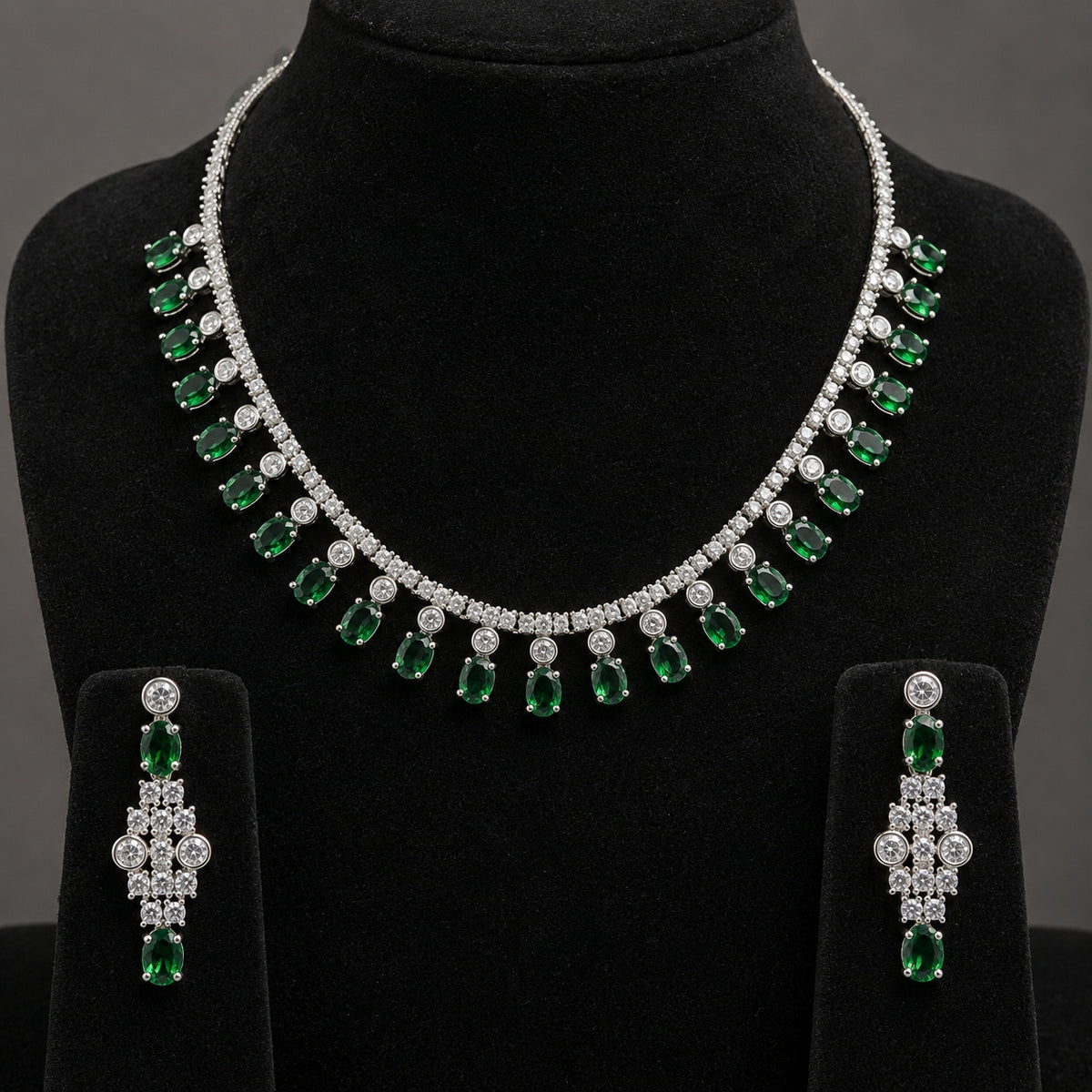 A silver-tone jewelry set featuring a collar necklace and matching drop earrings, both adorned with clear stones and green emerald-cut gemstones, displayed on a black velvet jewelry bust.