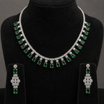 A silver-tone jewelry set featuring a collar necklace and matching drop earrings, both adorned with clear stones and green emerald-cut gemstones, displayed on a black velvet jewelry bust.