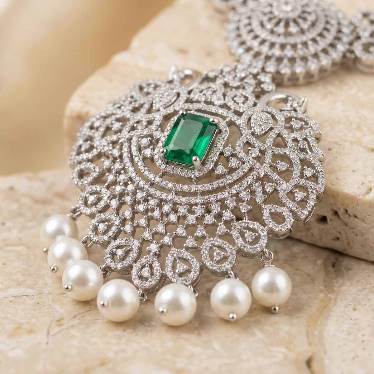 Silver-toned ornate pendant necklace with a rectangular green emerald-cut center stone and dangling white pearl beads, displayed on a beige stone surface.