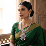A woman wearing a rich green saree with gold zari borders, styled with an elaborate silver-toned statement necklace and matching earrings, set against a warm, traditional architectural background.