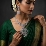 A woman wearing an ornate silver-toned jewelry set with green stone accents, including a necklace, earrings, and a bindi, paired with a dark green saree with gold borders against a dark gray background.
