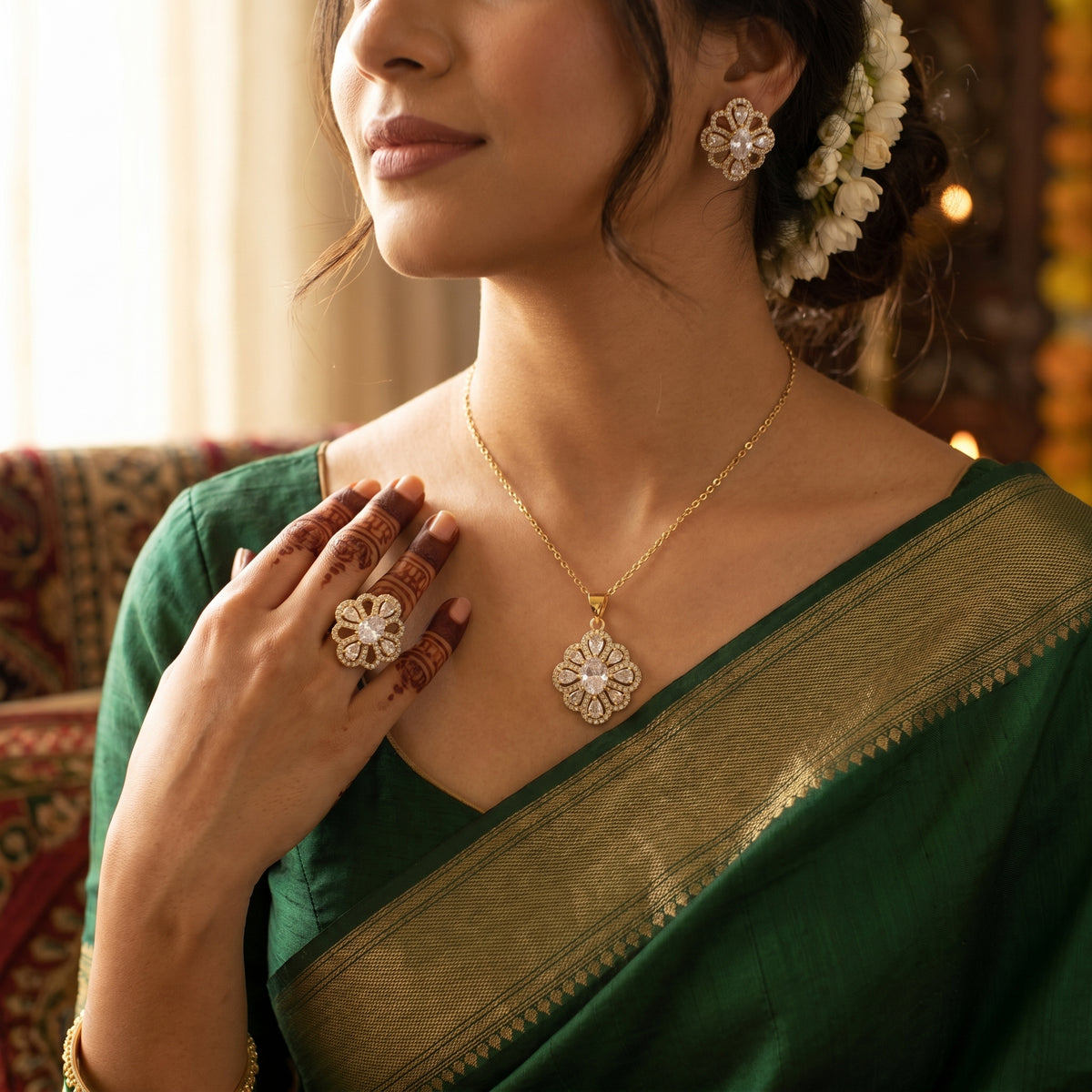 A woman wearing a gold pendant necklace and matching flower-shaped earrings, styled with a green and gold sari and floral hair accessories in a warm indoor setting.