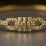 A gold-toned bangle bracelet featuring a central bowtie-like motif paved with small clear stones, shown in a close-up studio shot on a dark reflective surface.