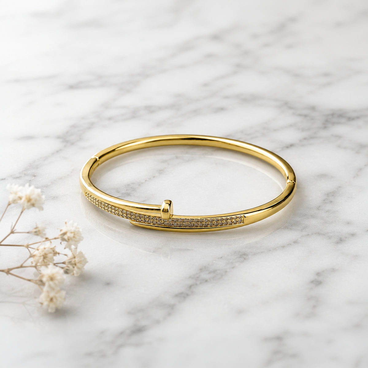 A polished gold bangle bracelet with a row of small clear stones along one side, displayed on a white marble surface with small white flowers nearby.