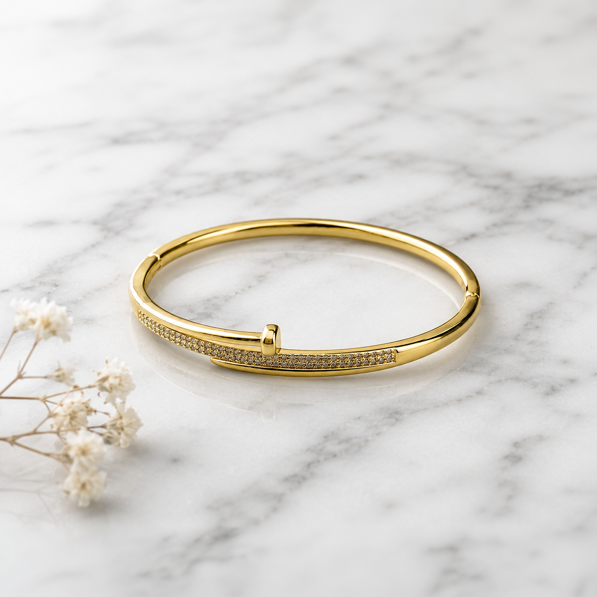 A polished gold bangle bracelet with a row of small clear stones along one side, displayed on a white marble surface with small white flowers nearby.