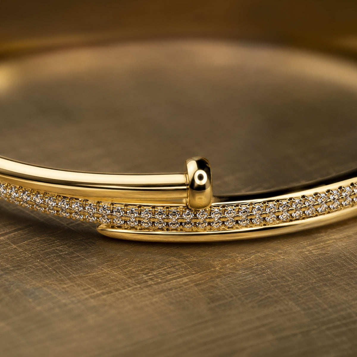 A close-up of a polished gold bangle bracelet with a double row of pavé-set clear stones, resting on a textured gold surface.