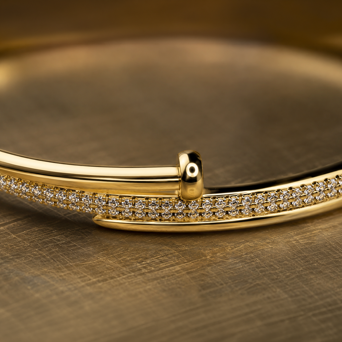 A close-up of a polished gold bangle bracelet featuring a double row of pavé-set clear stones along the front band, resting on a textured gold surface.