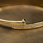 A close-up of a polished gold bangle bracelet with a double row of pavé-set clear stones, resting on a textured gold surface.