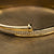 A close-up of a polished gold bangle bracelet with a double row of pavé-set clear stones, resting on a textured gold surface.