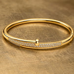 A polished gold bangle bracelet with a row of pavé-set clear stones across the front, displayed on a textured gold surface.