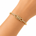 A gold-tone bangle bracelet with pavé-set clear stones and a central interlocking monogram design, shown worn on a person's wrist against a clean white background.
