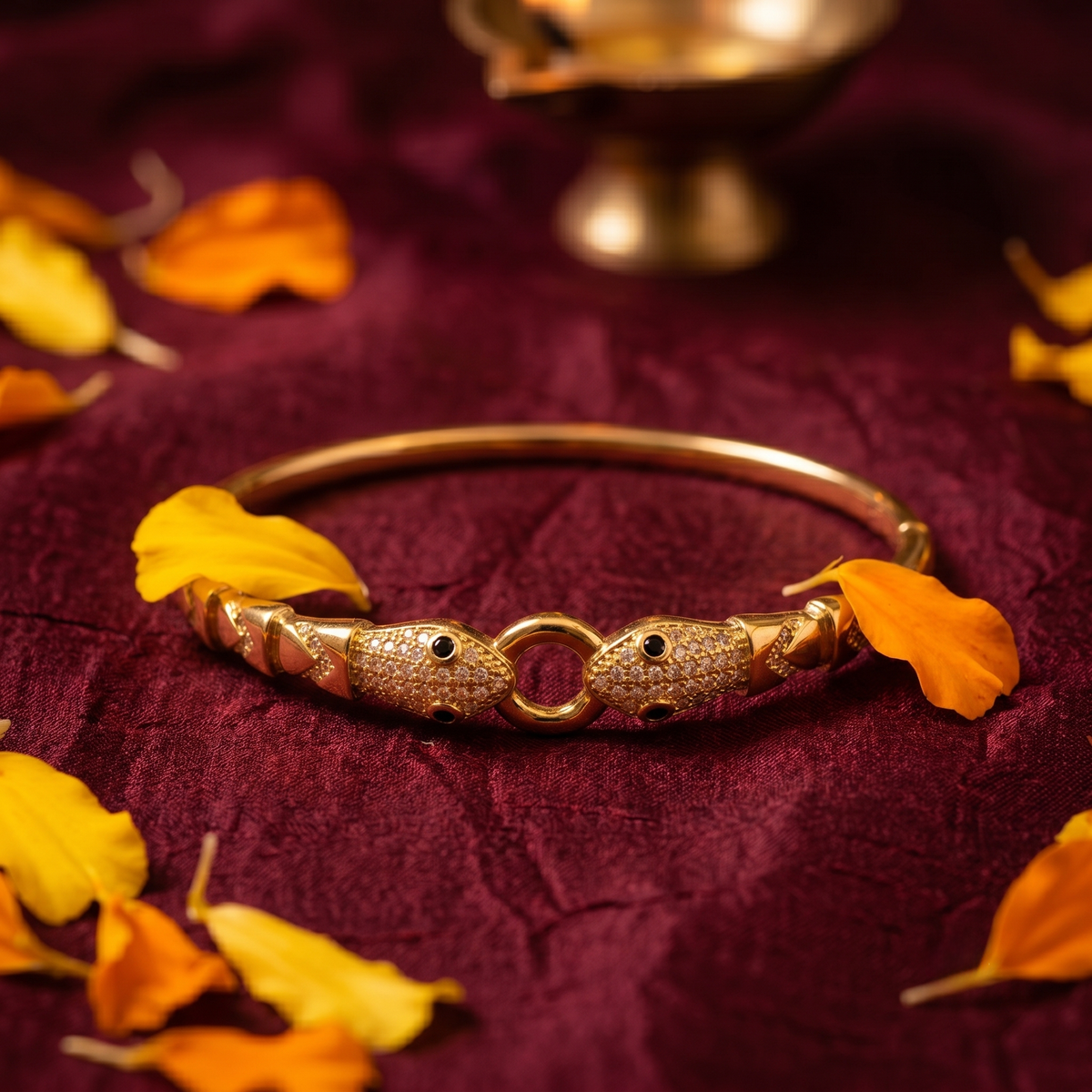 A gold-plated bangle bracelet with two fish-shaped charms encrusted with small clear stones, resting on a deep burgundy fabric backdrop scattered with orange and yellow leaves.