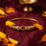 A gold-plated bangle bracelet with two fish-shaped charms encrusted with small clear stones, resting on a deep burgundy fabric backdrop scattered with orange and yellow leaves.