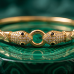 A gold bangle bracelet with two snake-head clasps paved with small clear stones and accented with black inlays, resting on a green marbled surface.