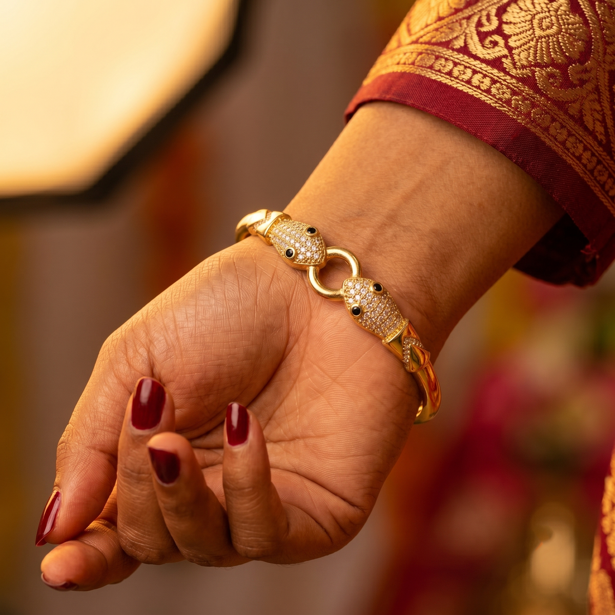 A close-up of a person's hand wearing a gold-plated snake-link bracelet adorned with pavé-set clear stones and small black bead eyes, paired with a deep red and gold embroidered sleeve.