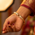 A close-up of a person's hand wearing a gold-plated snake-link bracelet adorned with pavé-set clear stones and small black bead eyes, paired with a deep red and gold embroidered sleeve.