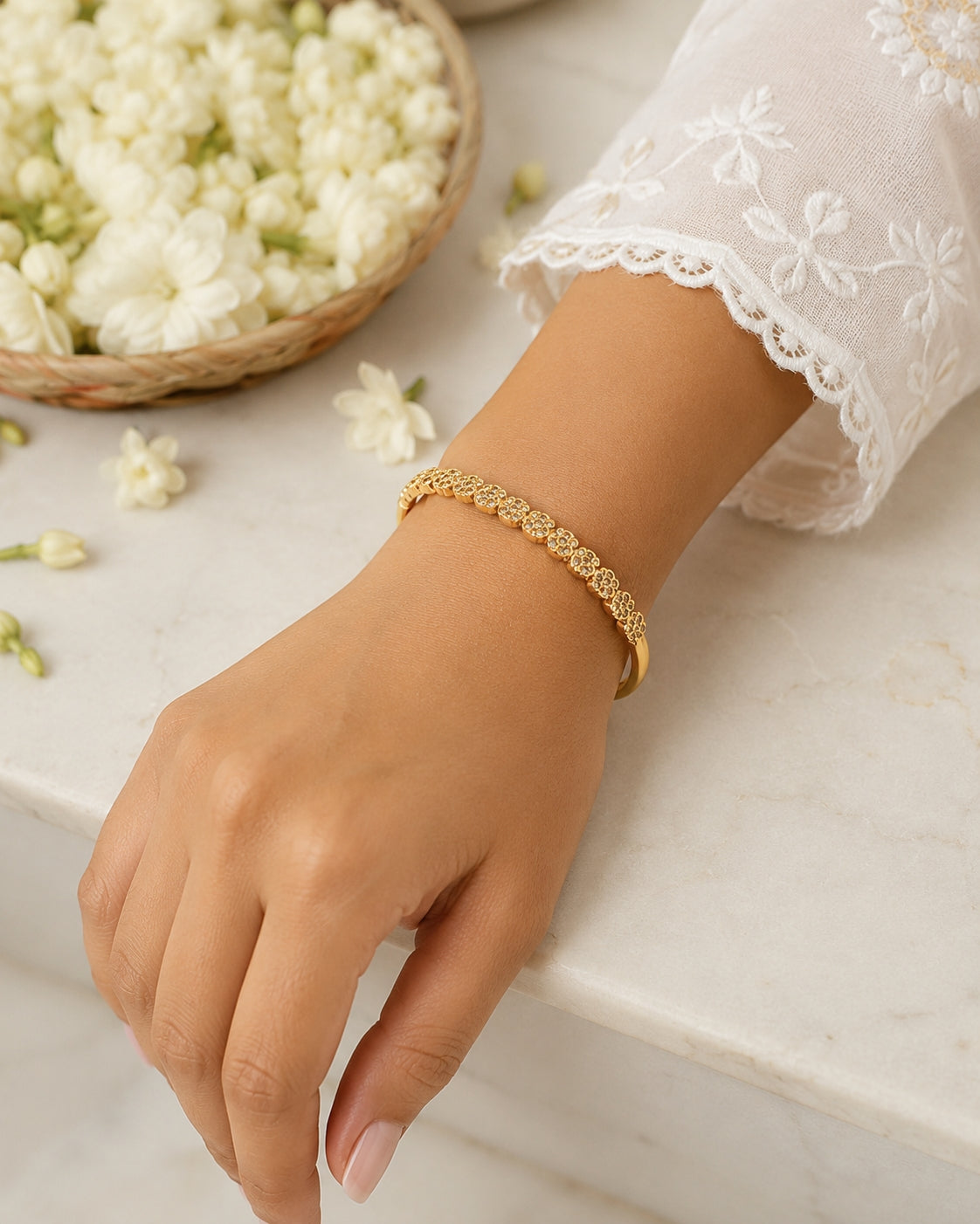 A delicate gold bracelet with a repeating floral pattern worn on a woman's wrist, shown in a styled lifestyle setting with white lace sleeves and a basket of white flowers.