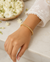 A delicate gold chain bracelet with small white floral charms, worn on a woman's wrist. The woman is wearing a white lace-sleeved blouse, and a woven basket filled with white flowers is visible in the background on a light marble surface.