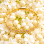 A gold bangle bracelet with a row of sparkling clear stones along the front, displayed on a bed of white jasmine flowers.
