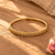 A gold bangle bracelet with a textured, floral-like pattern of small clear stones, displayed on a warm wooden surface with soft shadows.
