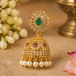 An ornate gold-plated jhumka earring with a green oval stone at the top, surrounded by clear stones, and a bell-shaped lower portion adorned with red stones, clear stones, and dangling white pearls, set against a warm neutral background with decorative flowers.