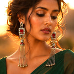 A close-up lifestyle shot of a woman wearing ornate silver-toned tassel earrings with rectangular red inlay stones, set against a warm golden sunset background.