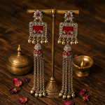 A pair of ornate silver-toned Indian jhumka earrings with red inlay and dangling chain tassels, displayed on a brass stand over a dark wooden surface with decorative brass vessels and scattered rose petals.