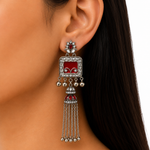 A close-up of a woman's ear wearing a large silver-toned tassel earring with a red enamel center panel, rhinestone detailing, and dangling silver bells and chains.