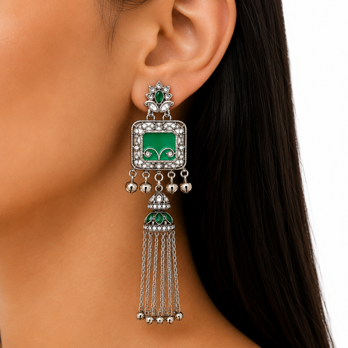 A close-up of a woman's ear wearing a large silver-toned dangle earring with green enamel details and small clear stones.