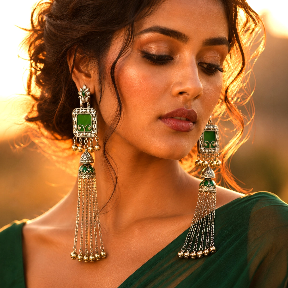 A woman wearing large green and silver tassel earrings, shown in a close-up portrait with warm golden sunlight and a blurred outdoor background.