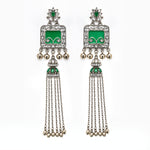 A pair of traditional Indian silver-toned dangle earrings with vibrant green enamel panels, clear pavé stones, and dangling silver chains ending in small gold-toned bells, shown against a plain white background.