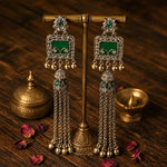A pair of ornate silver-toned tassel earrings with rectangular green stone inlays, displayed on a brass stand over a dark wooden surface with small brass vessels and scattered rose petals.